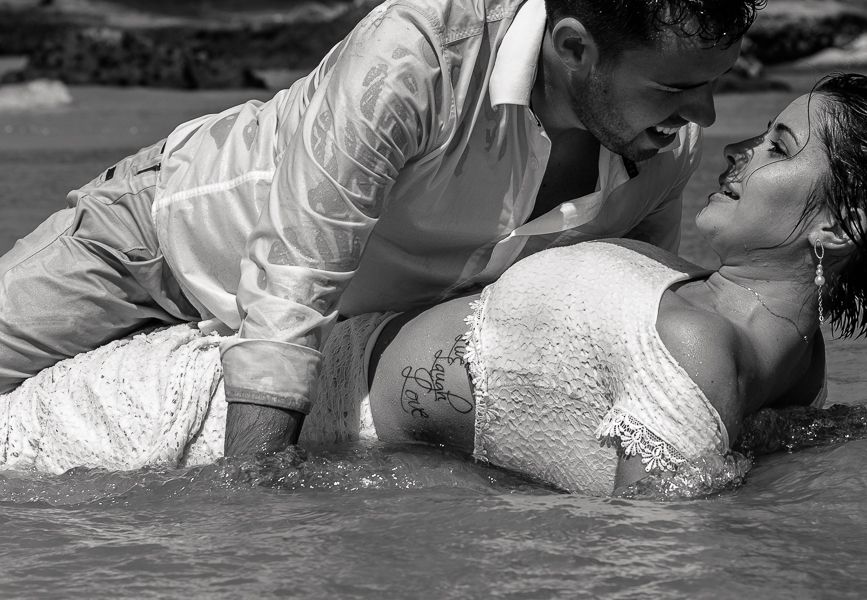 Black and white photo of a romantic couple in wedding attire laying in shallow water during a photo shoot.