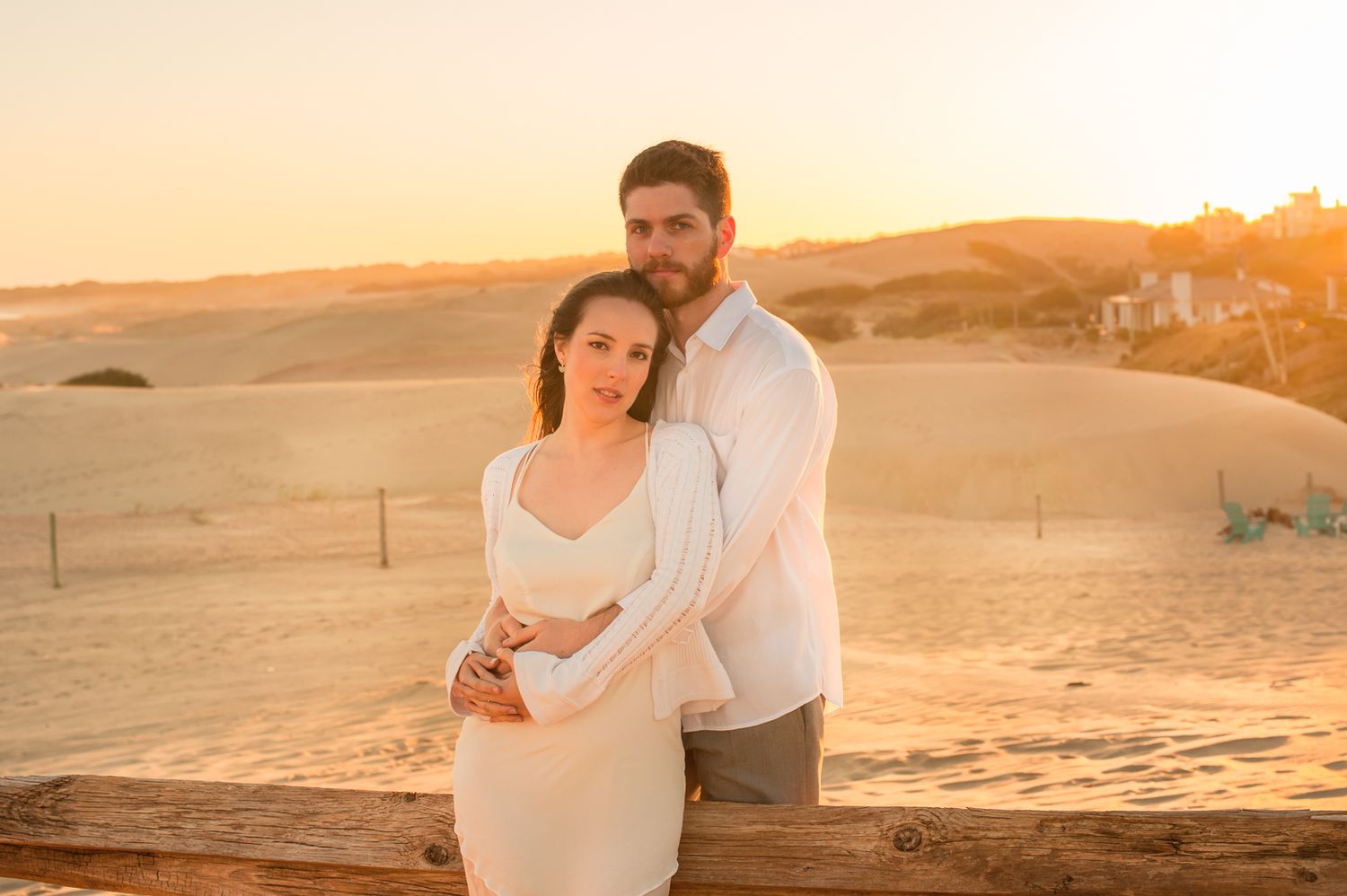 A couple embraces during golden hour sunset at a scenic desert location with sand dunes in the background.