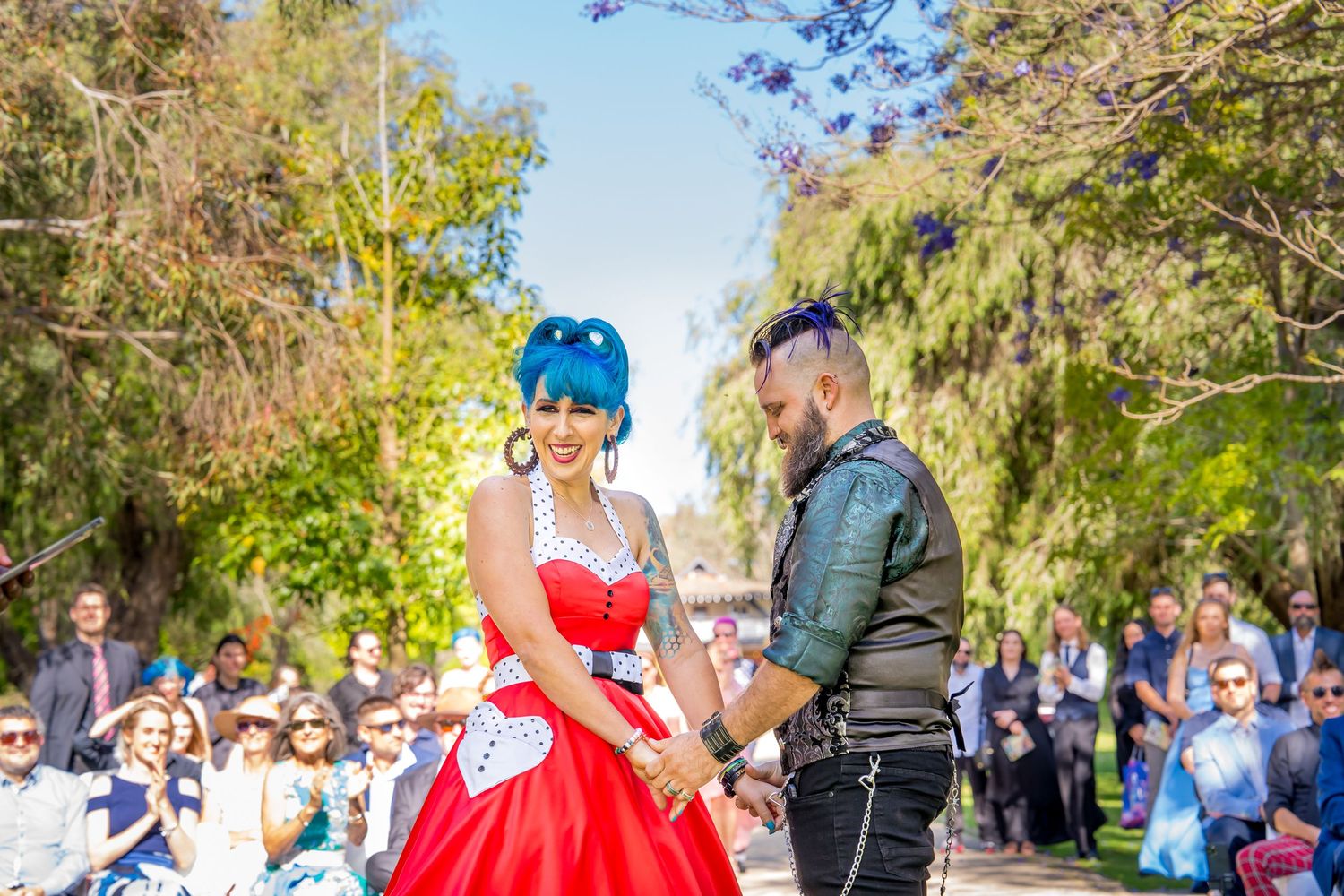 A couple dancing outdoors in a park setting with spectators watching, woman wearing a vibrant red dress.