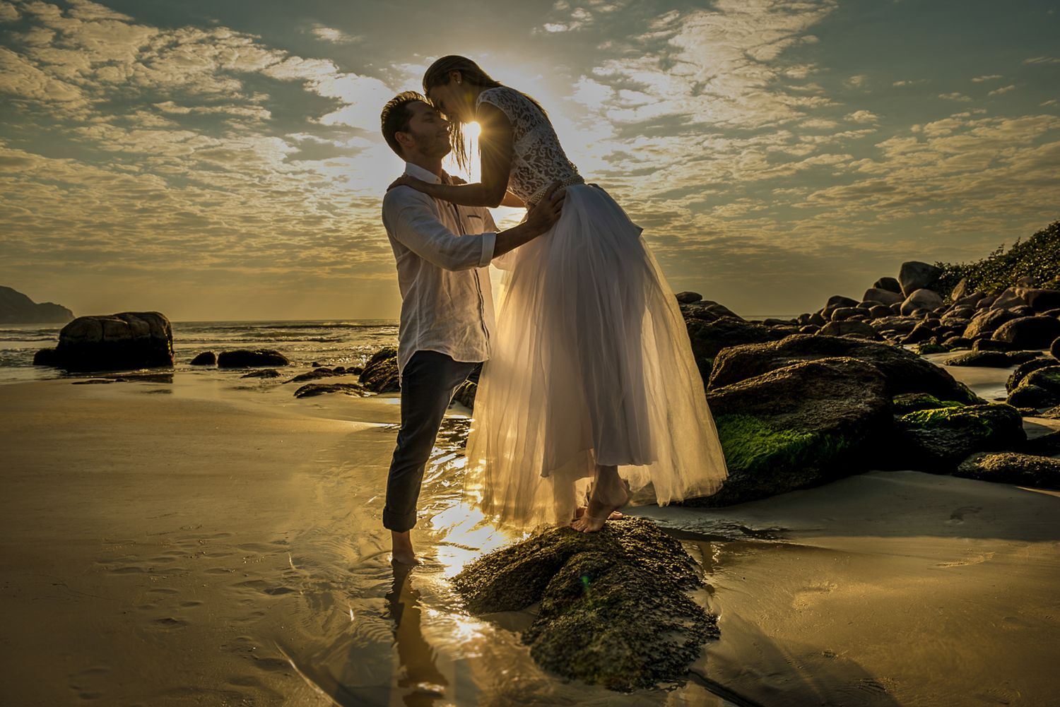 A romantic silhouette of a couple embracing on a beach at sunset with dramatic clouds and rocky shoreline.