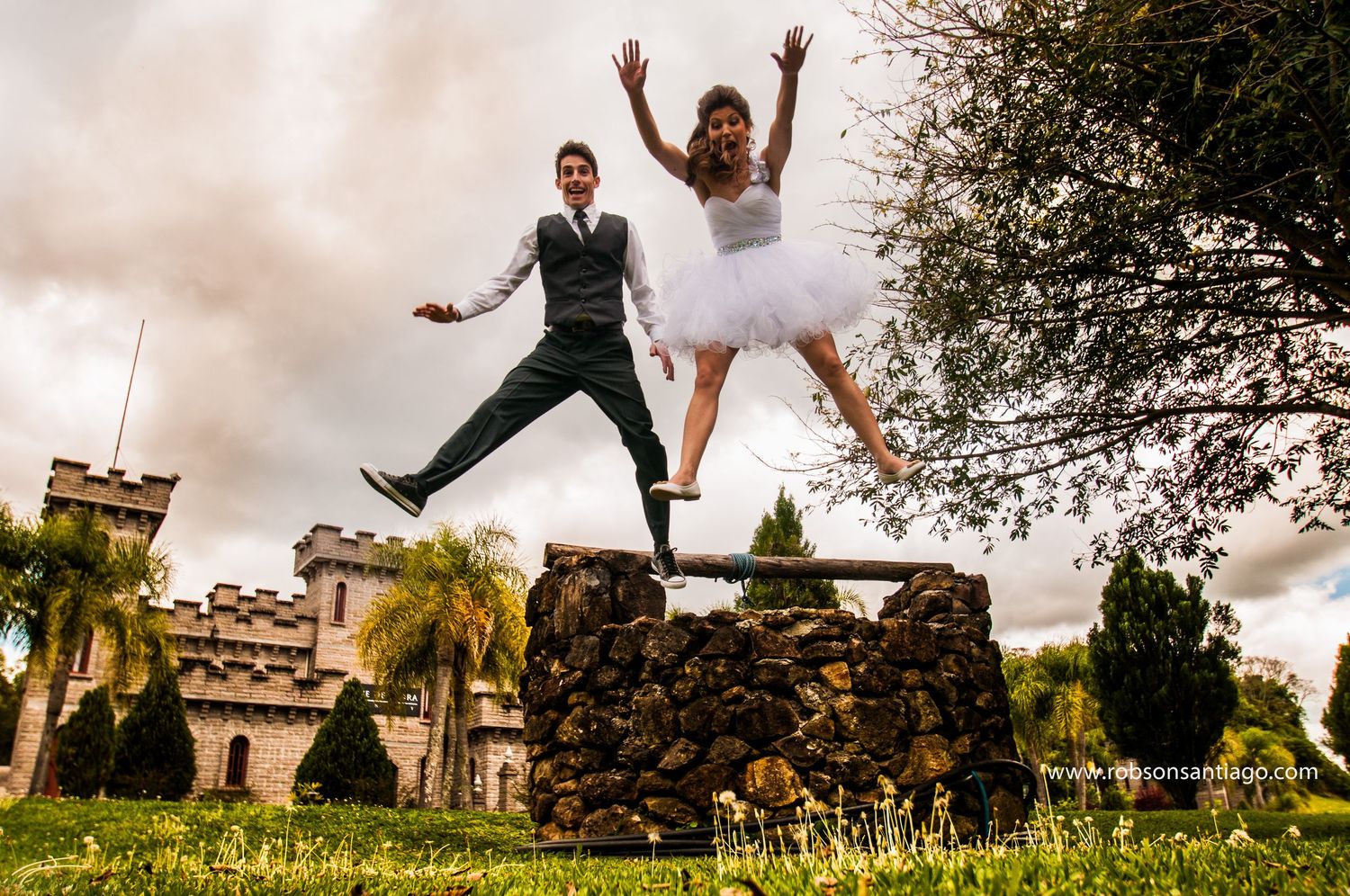 A joyful couple in wedding attire leaping together from a stone wall in a garden setting.