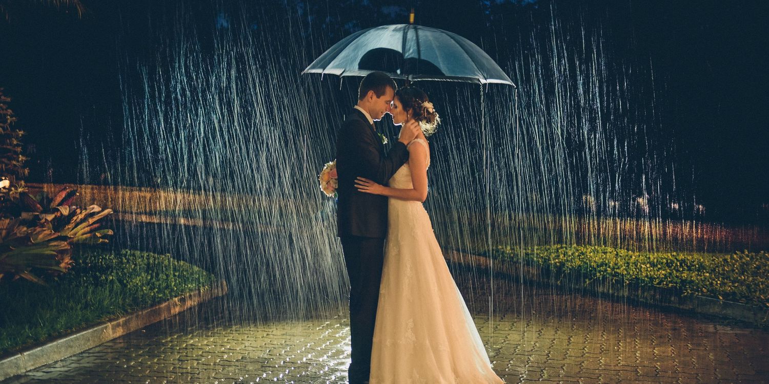 Couple in wedding attire share romantic moment under umbrella in rain at night with dramatic lighting.