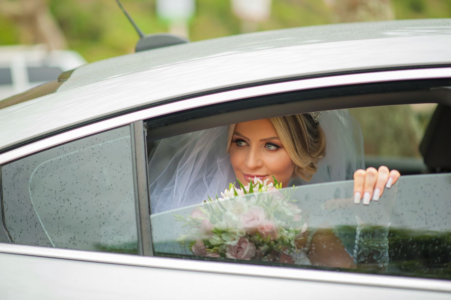 A bride smiles through a car window while holding a bouquet on her wedding day.