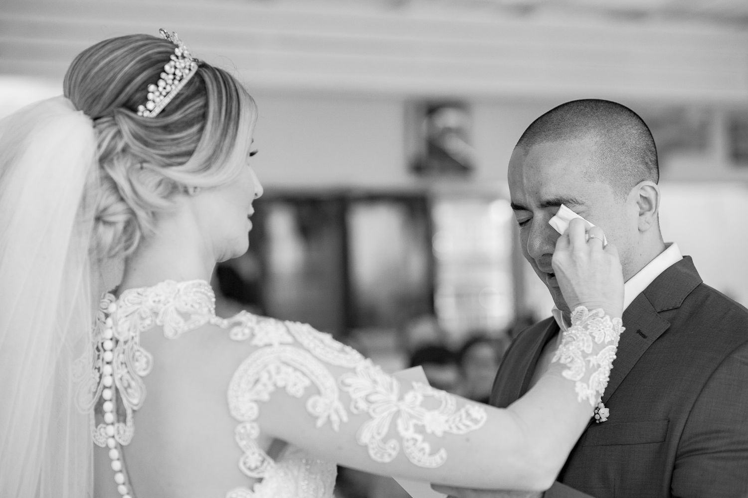 Emotional black and white candid moment at wedding ceremony with bride in lace gown wiping groom's tears.