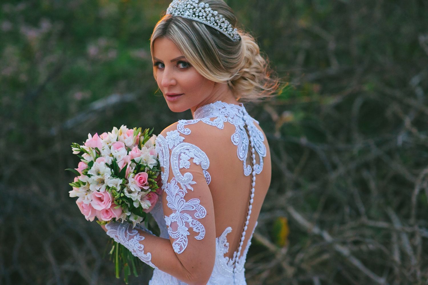 A bride in a backless lace wedding dress with long sleeves holds a pink bouquet while looking over her shoulder.