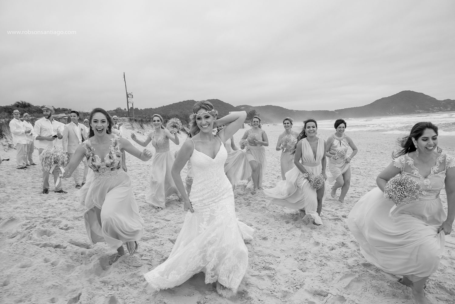 Bride and bridesmaids running on a beach in flowing white dresses with mountains in the background.