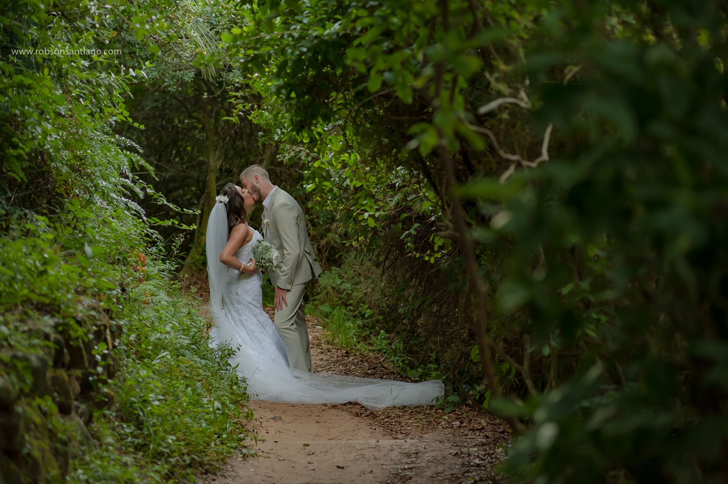 A romantic wedding couple embraces on a lush green forest path surrounded by natural foliage.