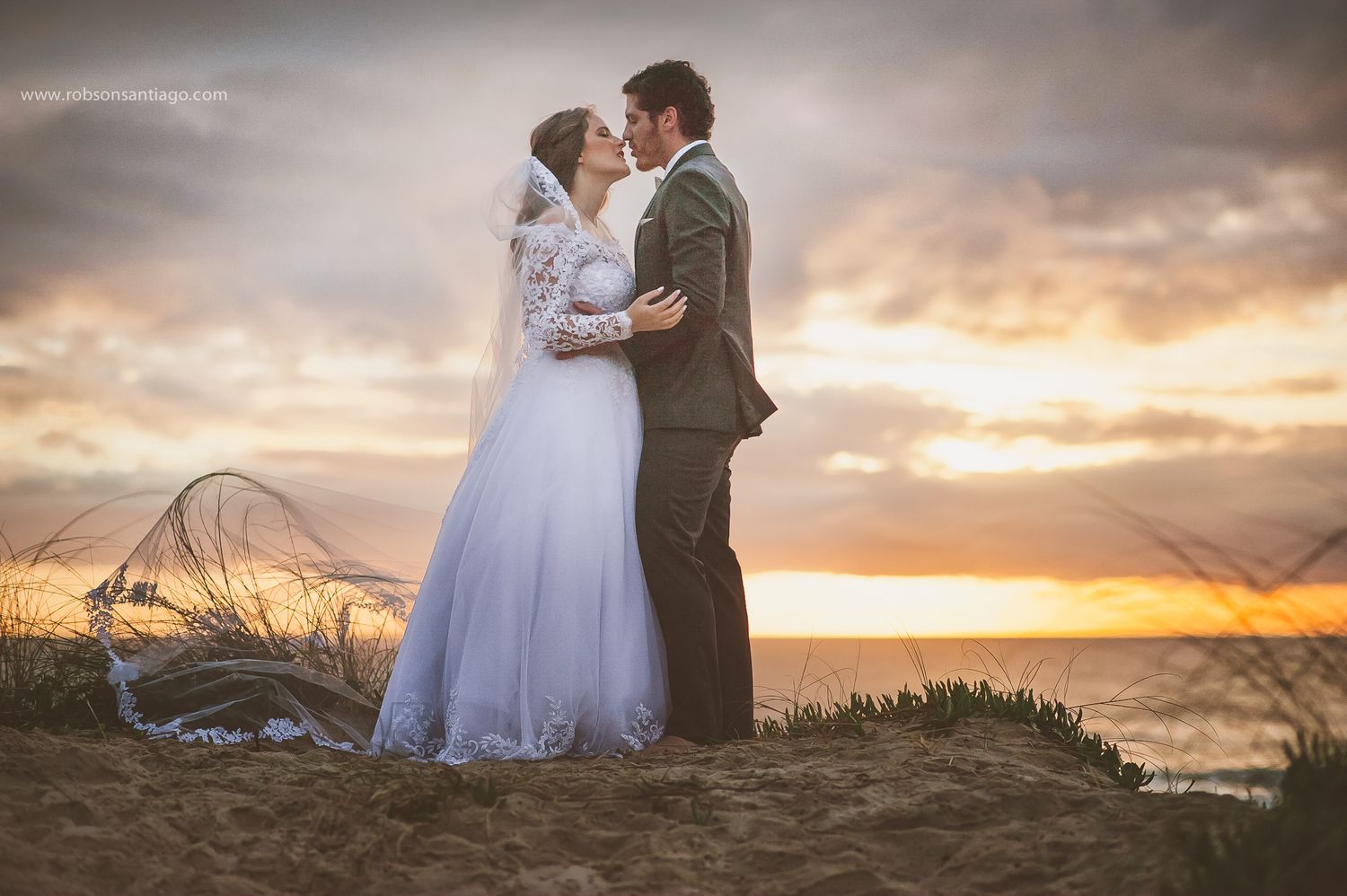 A couple in wedding attire share an intimate moment on a hilltop during sunset as a veil flows in the wind.