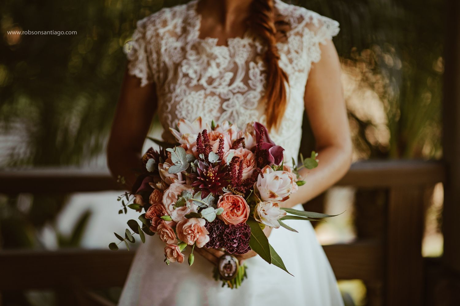 A bouquet of burgundy and blush roses is held against a white lace wedding dress in soft outdoor lighting.