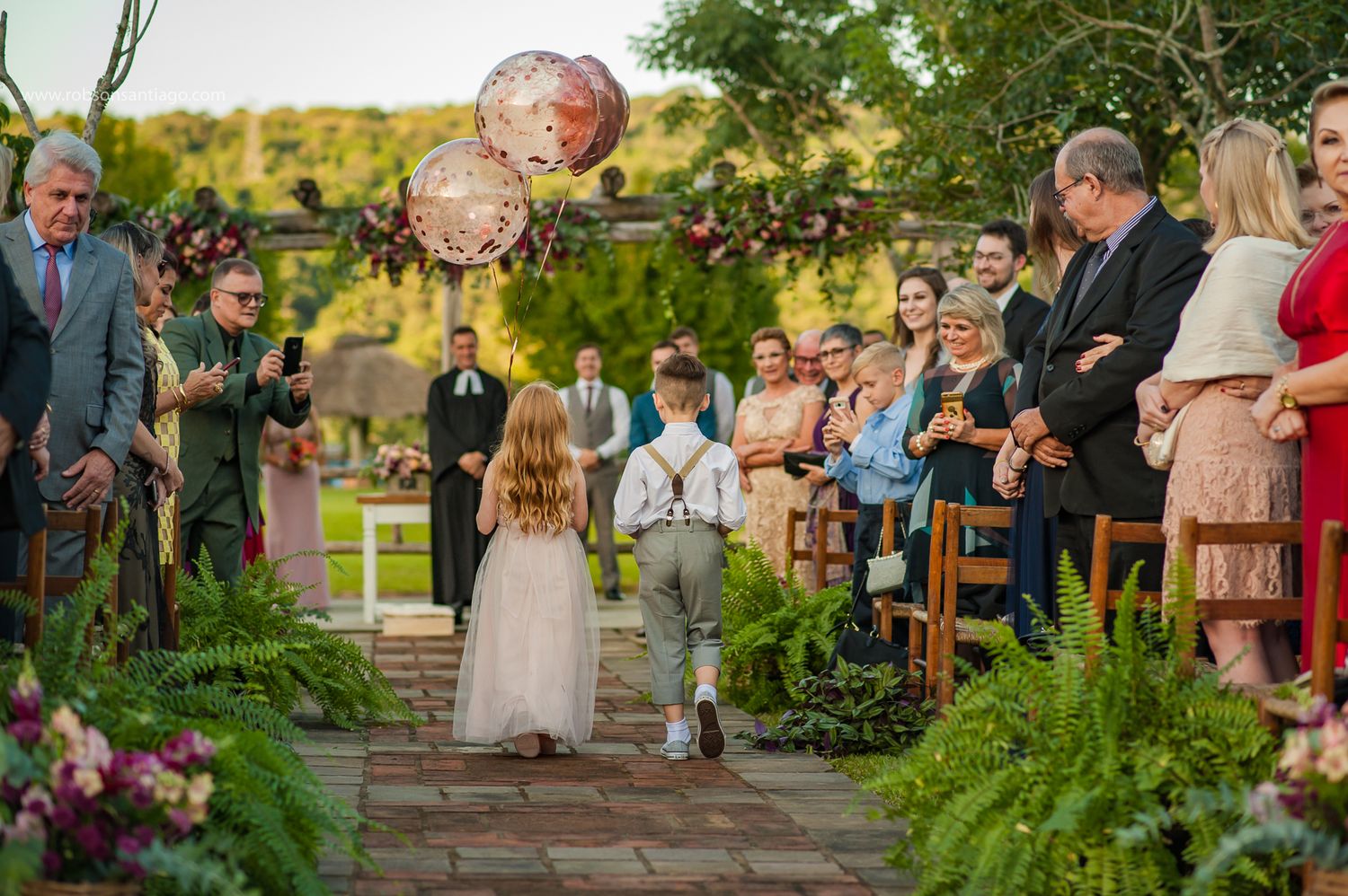 An outdoor garden wedding ceremony features a stone walkway lined with ferns and pink lanterns overhead in golden sunlight.