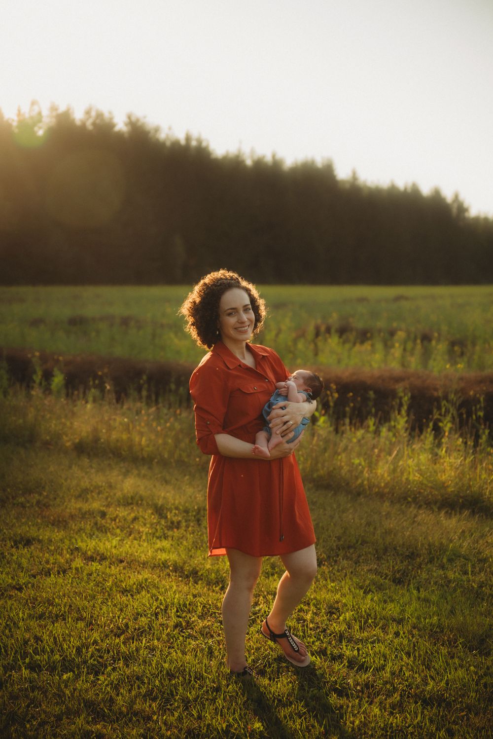 Figure in a bright red dress holds an infant while standing in a golden-lit field at sunset.