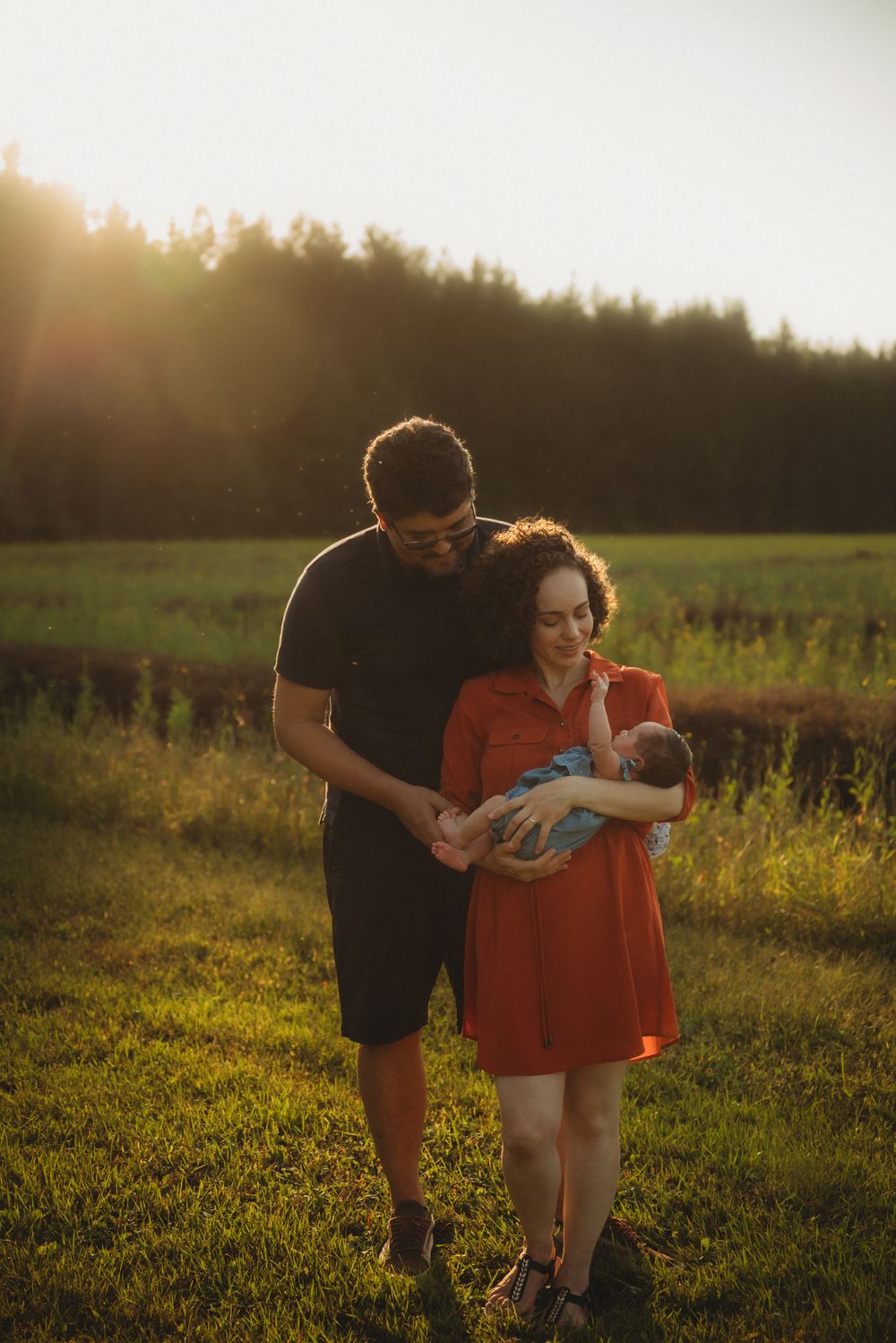 A couple embraces while holding their newborn in a grassy field during golden hour.