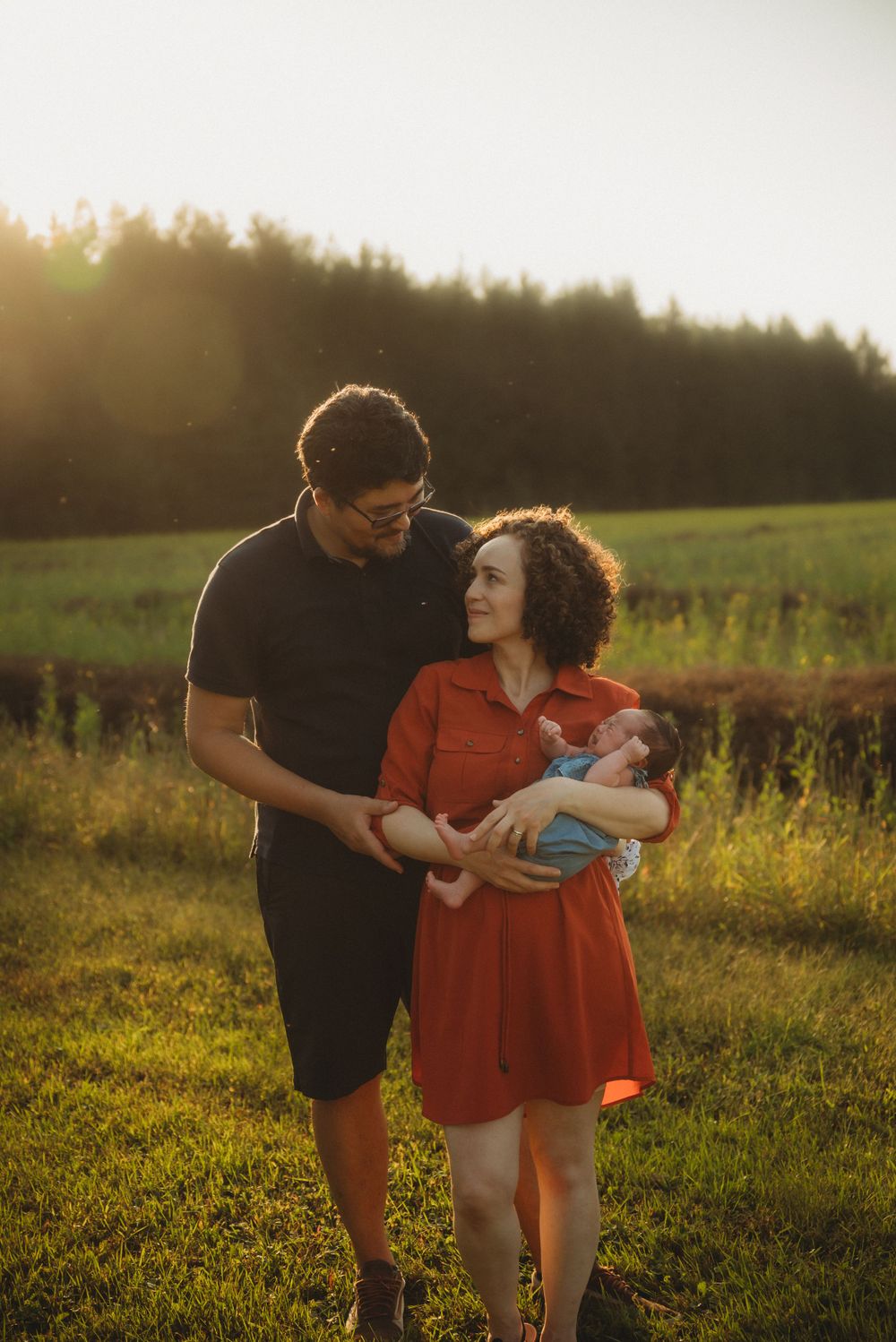 A tender family moment captured in a sun-drenched field with forest backdrop.