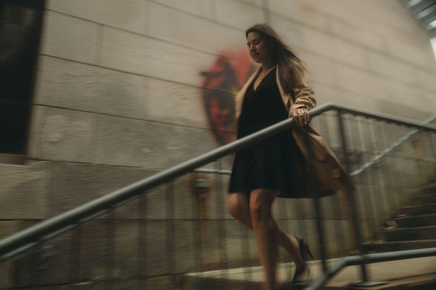Motion-blurred figure in black dress ascending stairs with handrail against neutral wall in artistic urban photography.