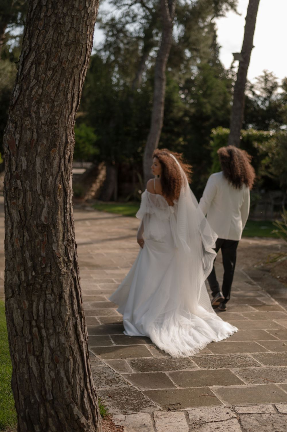 A wedding couple walks along a stone path surrounded by trees during golden hour.