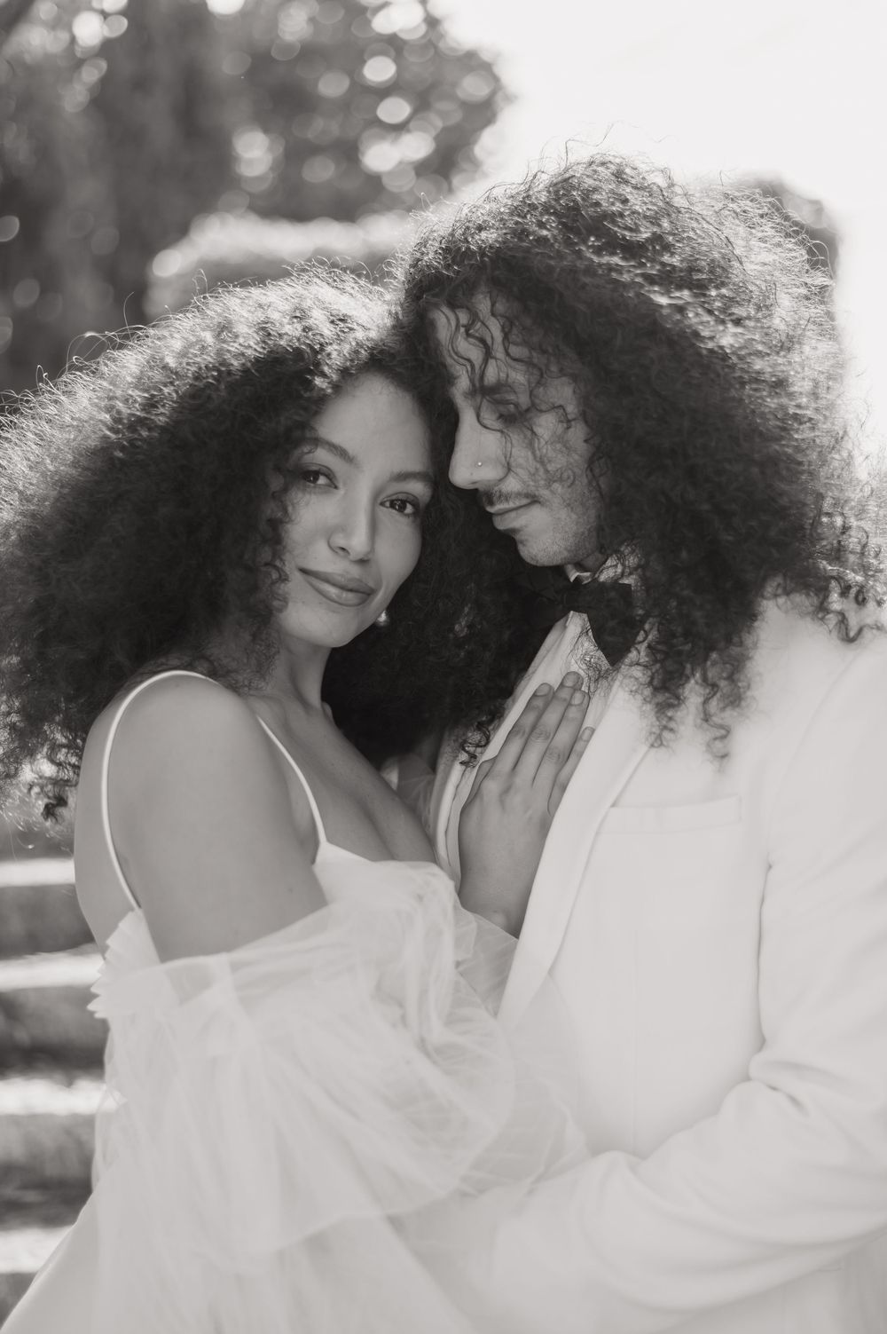 Romantic black and white portrait showing couple in white outfits with natural curly hair embracing outdoors.