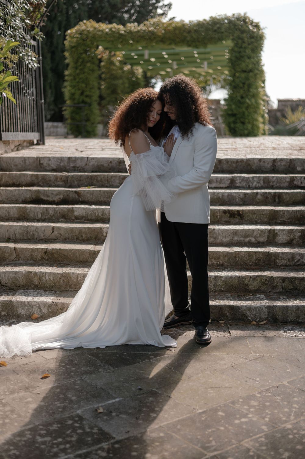 A romantic wedding couple embracing on stone steps with greenery in the background.