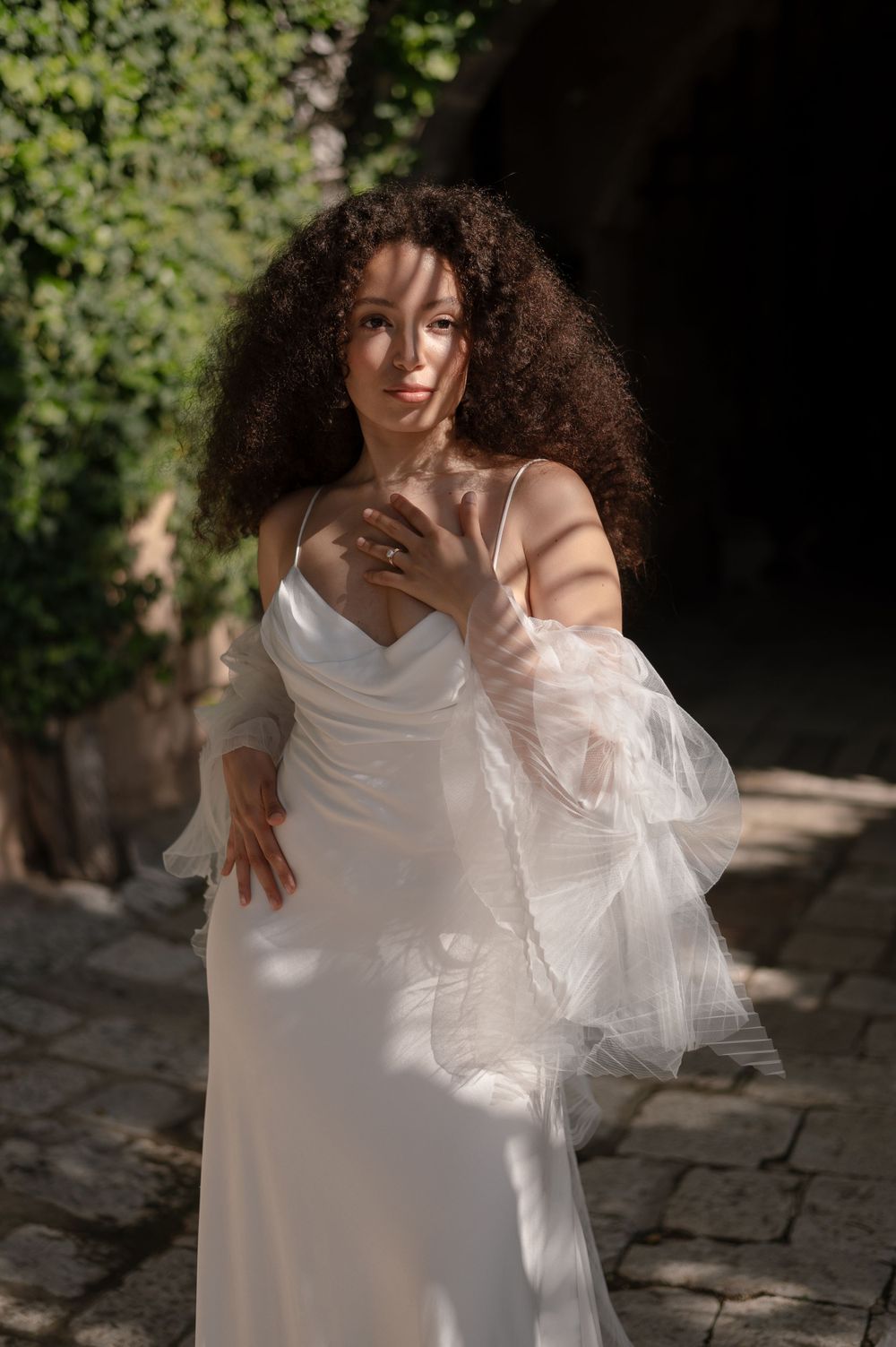 Elegant bride in white flowing dress with sheer sleeves poses outdoors with voluminous curly hair.