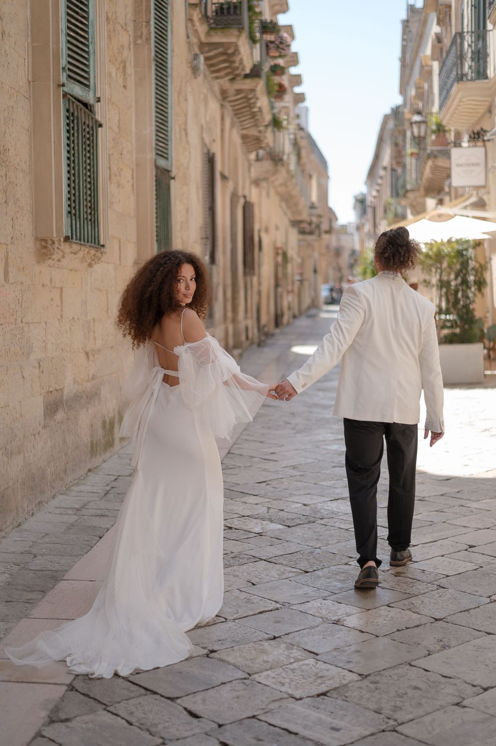 A couple in wedding attire walks hand in hand down a charming European stone street with historic buildings.