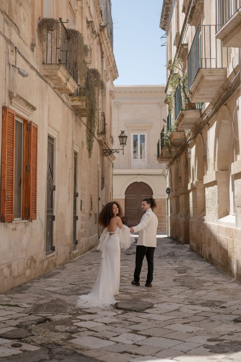 A romantic couple in white attire dancing in a charming Italian alleyway with historic stone buildings.