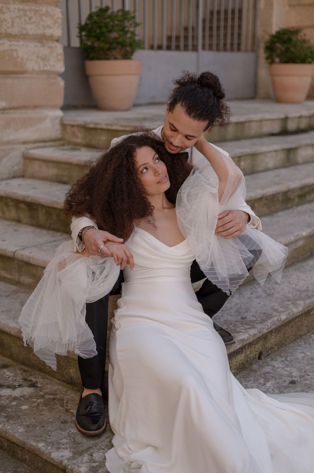 A romantic wedding couple sits together on stone steps with potted plants in the background.