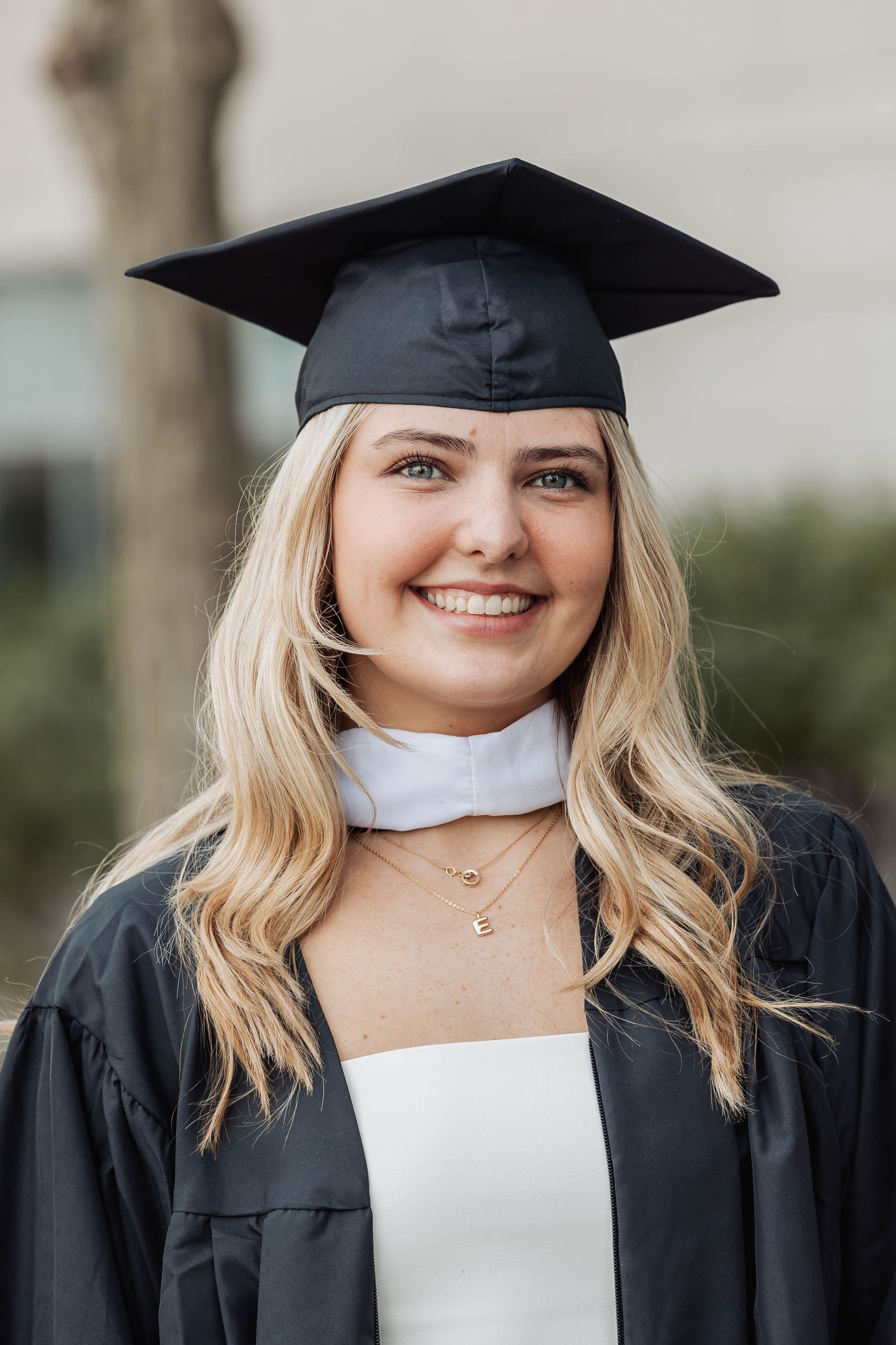 Graduate wearing black cap and gown with white dress smiles during commencement at university campus.