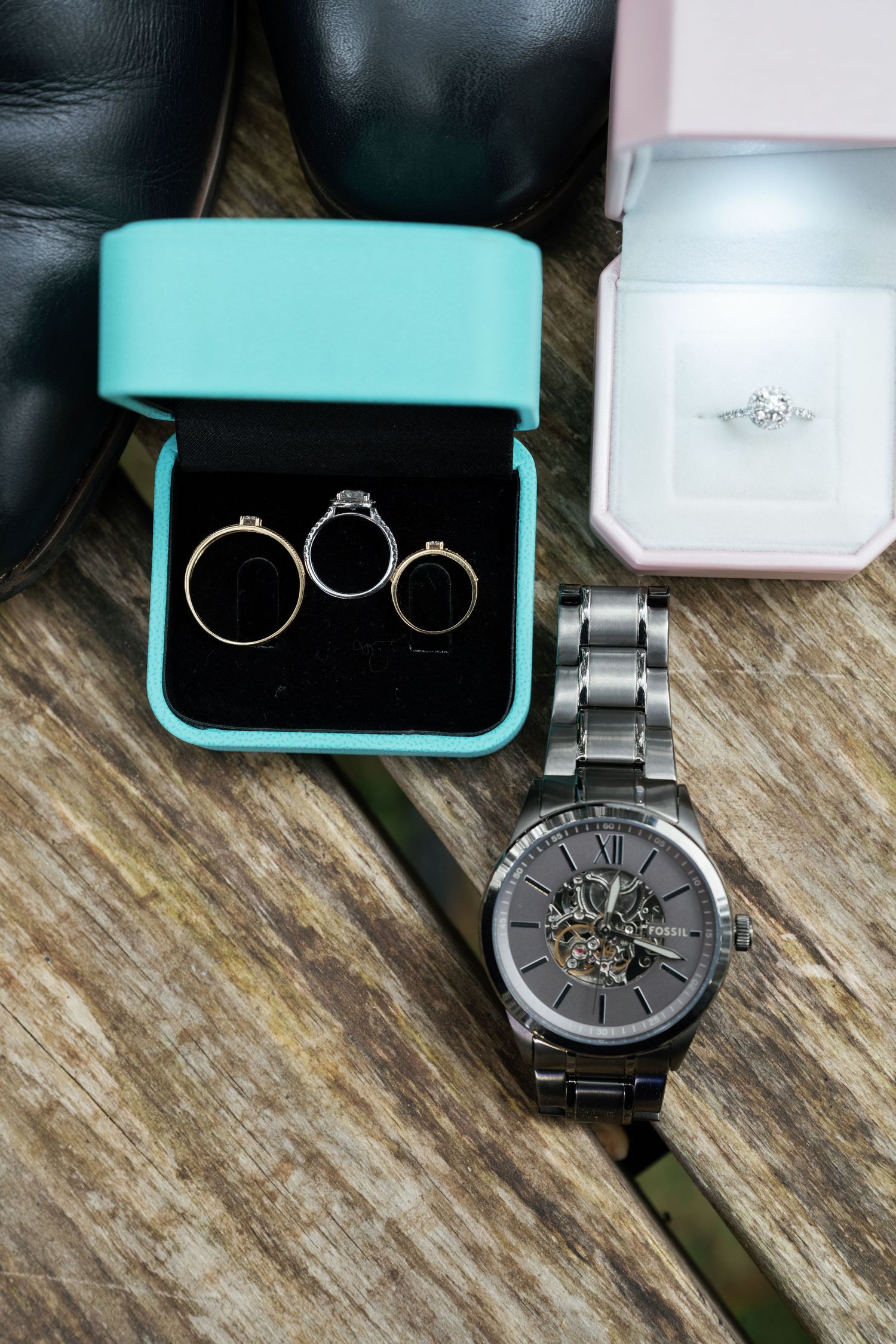 Three engagement rings in a turquoise box alongside a dark metal watch with exposed mechanical face on wooden surface.
