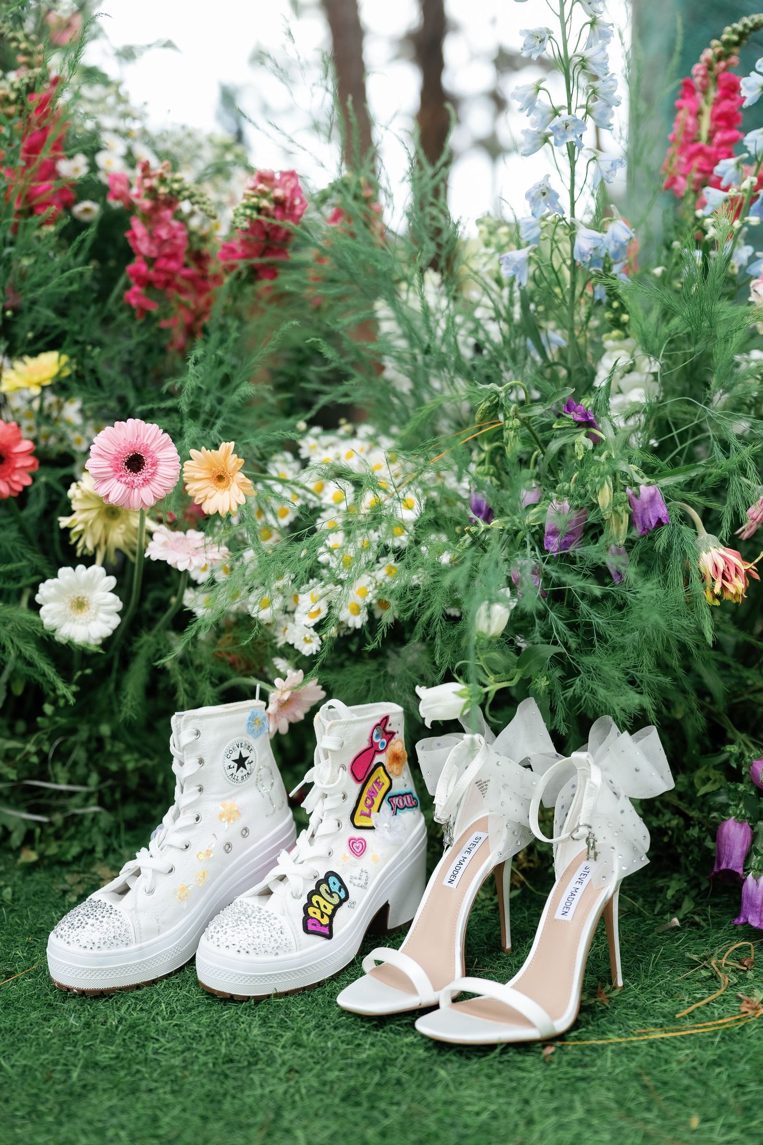A pair of decorated white high-top sneakers and white bow heels arranged on grass with colorful garden flowers in background.