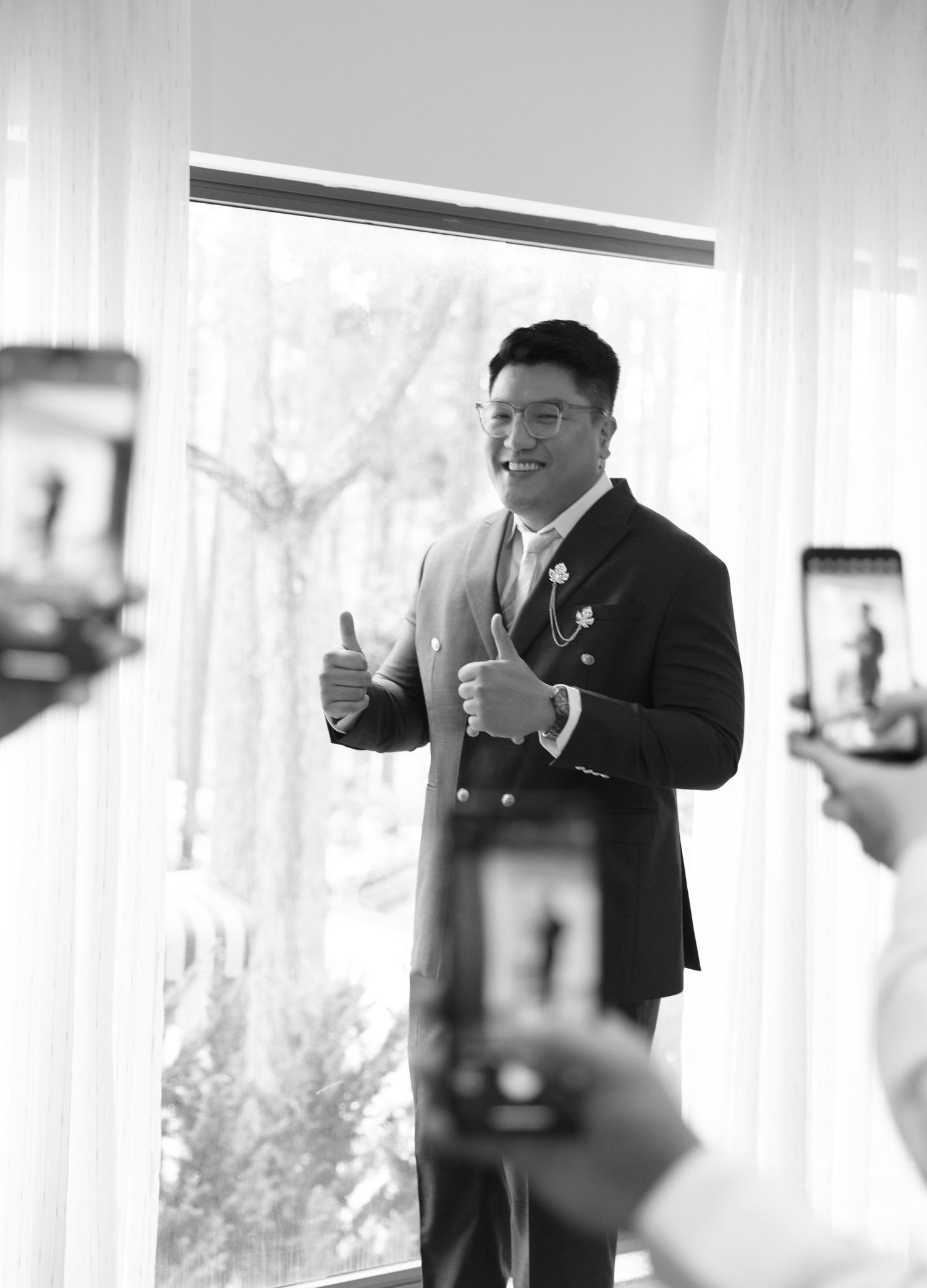 A groom in a dark suit gives enthusiastic thumbs up at a wedding venue, captured in black and white photography.