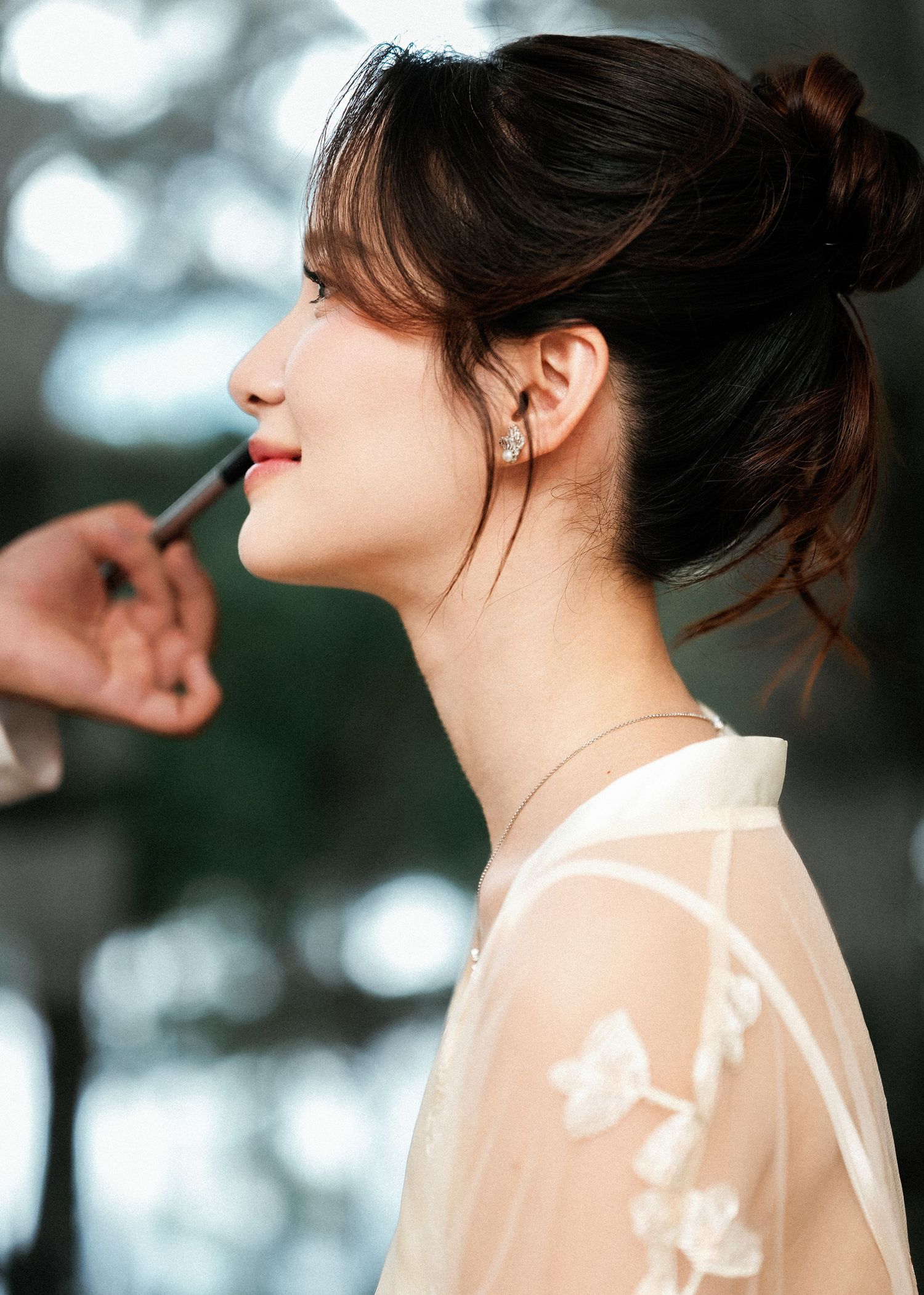Side profile portrait in soft lighting showing a woman with dark hair in a bun wearing a sheer white embroidered top.