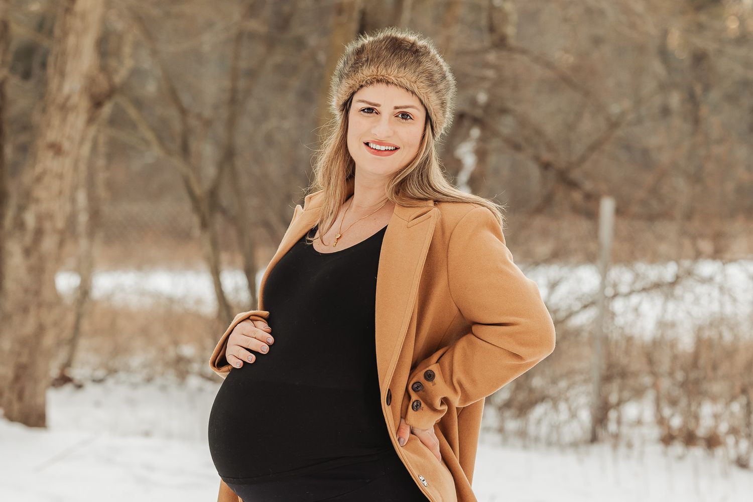 Pregnant woman wearing a tan beanie and camel coat poses outdoors in a snowy winter setting.