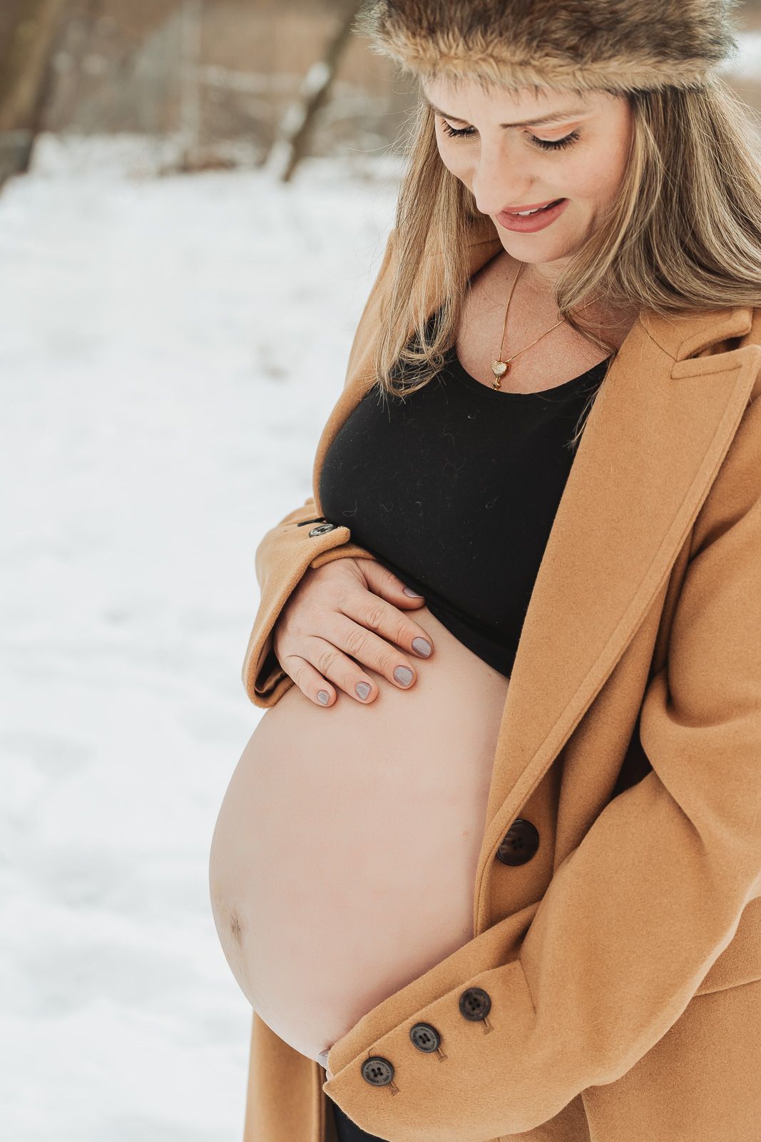 A pregnant woman in a tan winter coat and fur hat gently cradles her belly while standing in the snow.