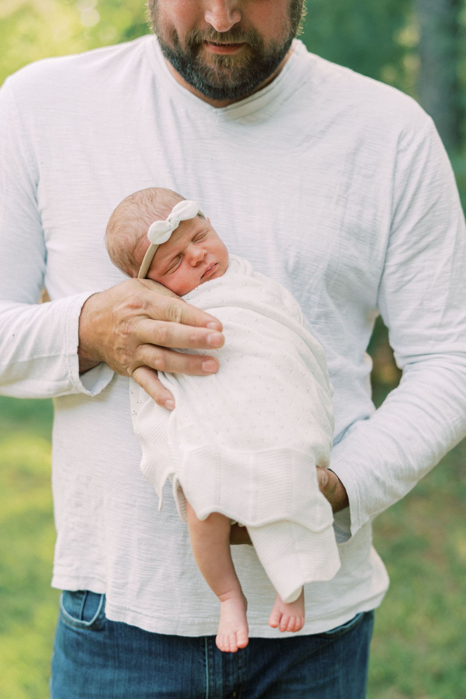 Parent in white shirt holds sleeping newborn wearing delicate headband outdoors.