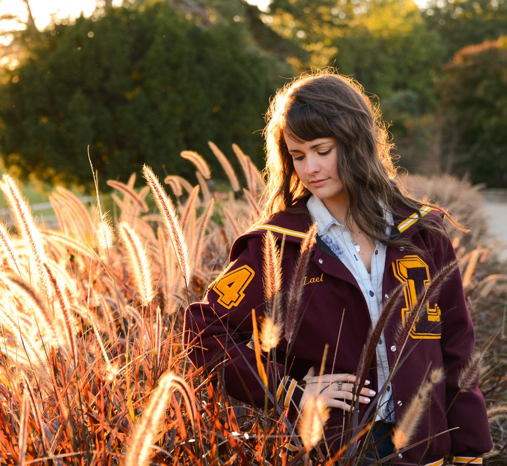 Person in burgundy letterman jacket stands among golden wheat at sunset.