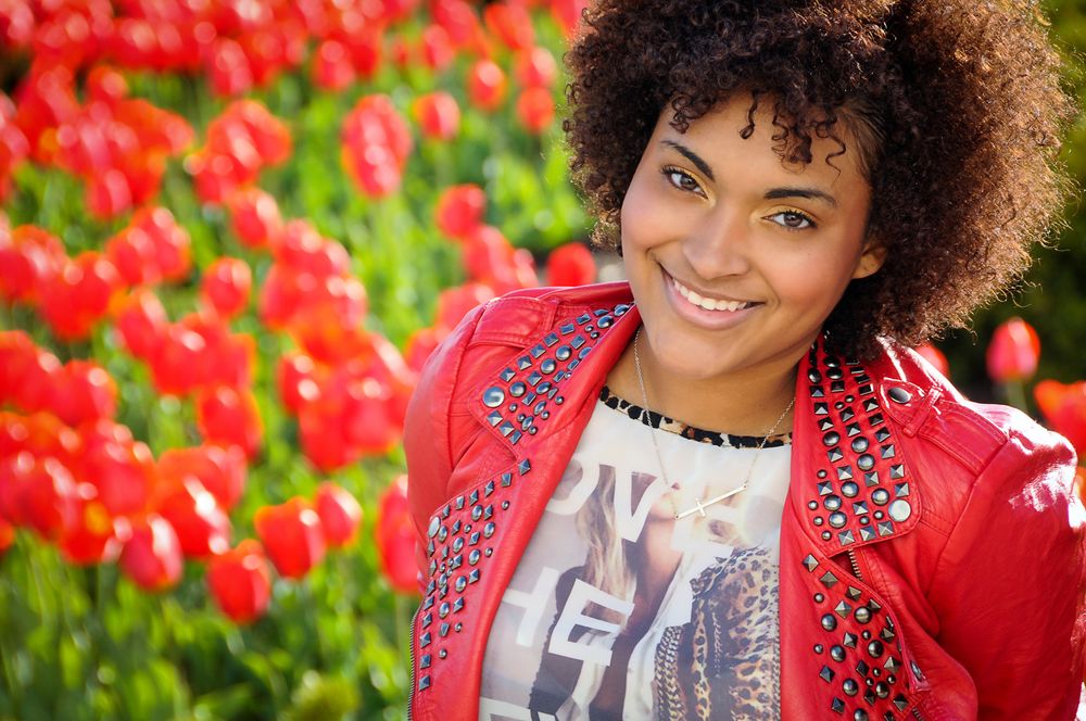 Portrait against red flowers wearing a vibrant red jacket and graphic t-shirt with natural curly hair.