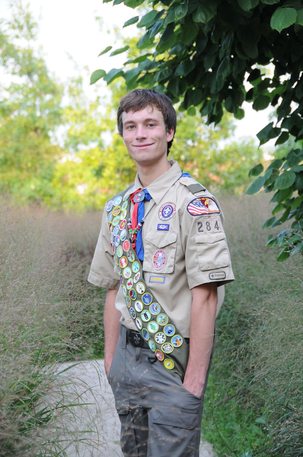 Boy Scout displaying uniform with numerous merit badges and patches while standing outdoors.