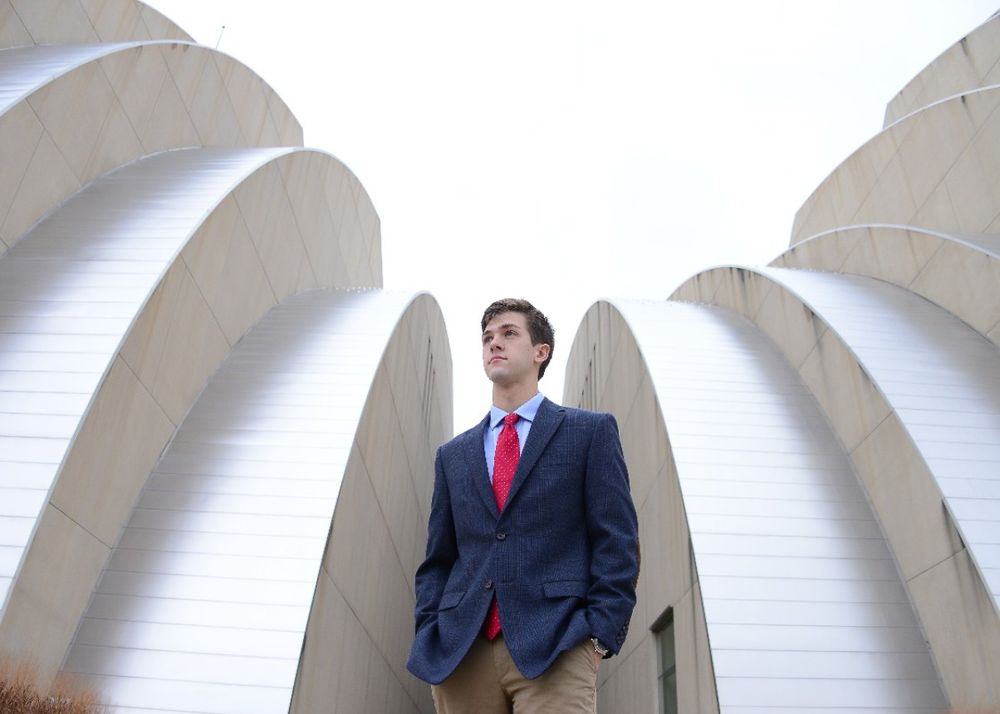 Figure in navy suit and red tie stands against white architectural arches.