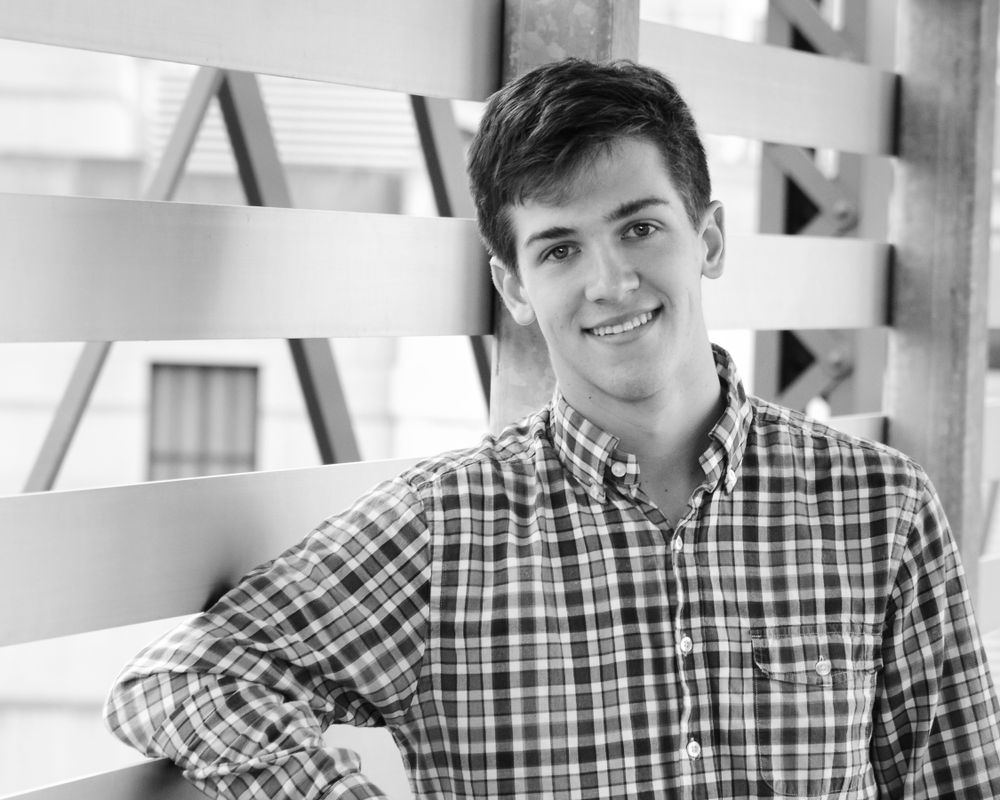 Black and white portrait of someone in a plaid button-down shirt leaning against a white fence.