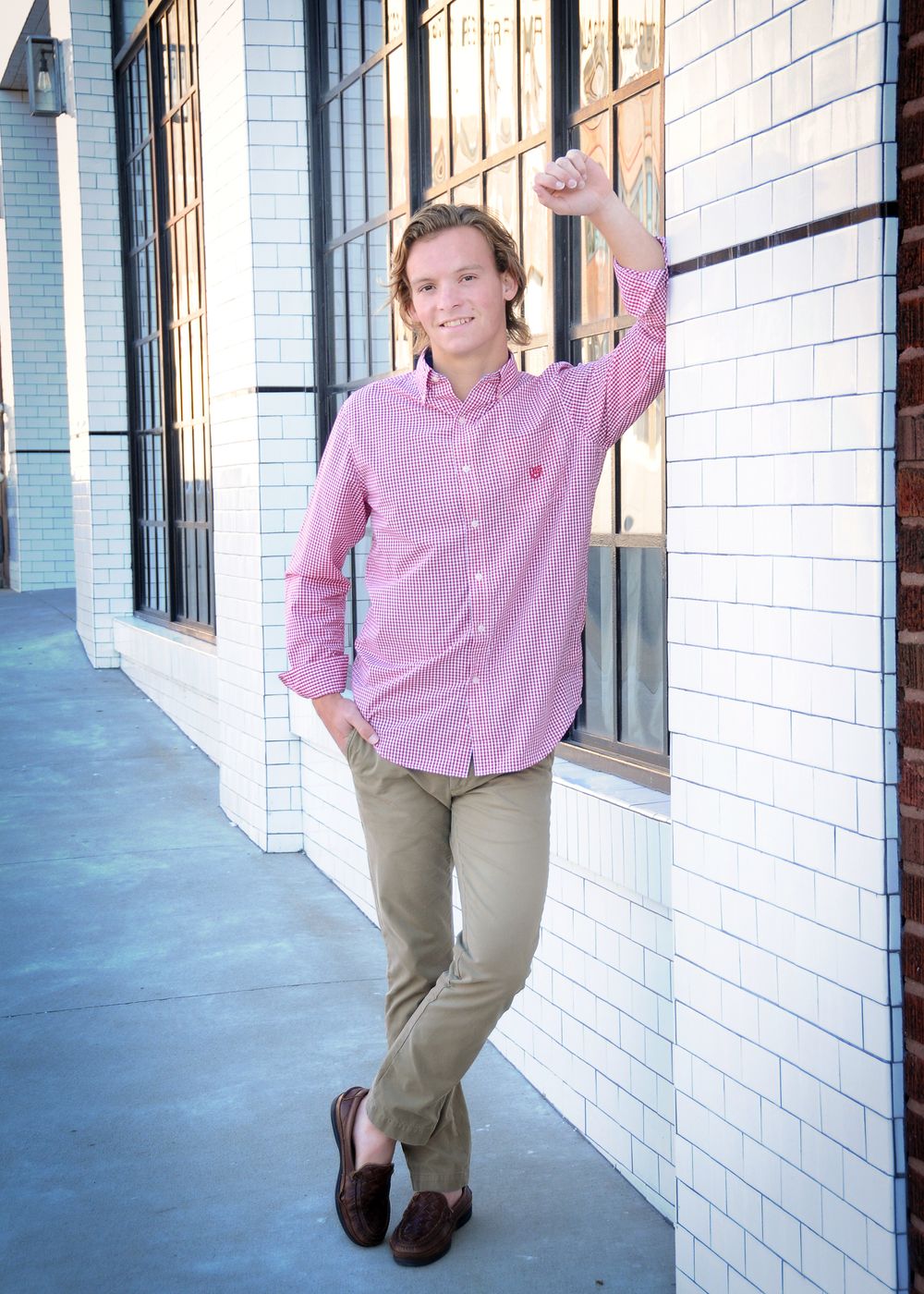 Casual pose against white brick wall wearing pink gingham shirt and khakis.