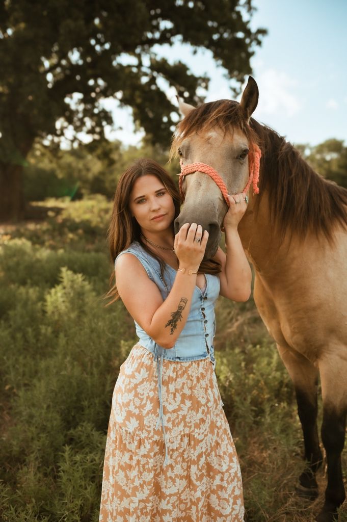 A woman in a floral dress and denim vest gently interacts with a brown horse on a sunny day in a grassy field.
