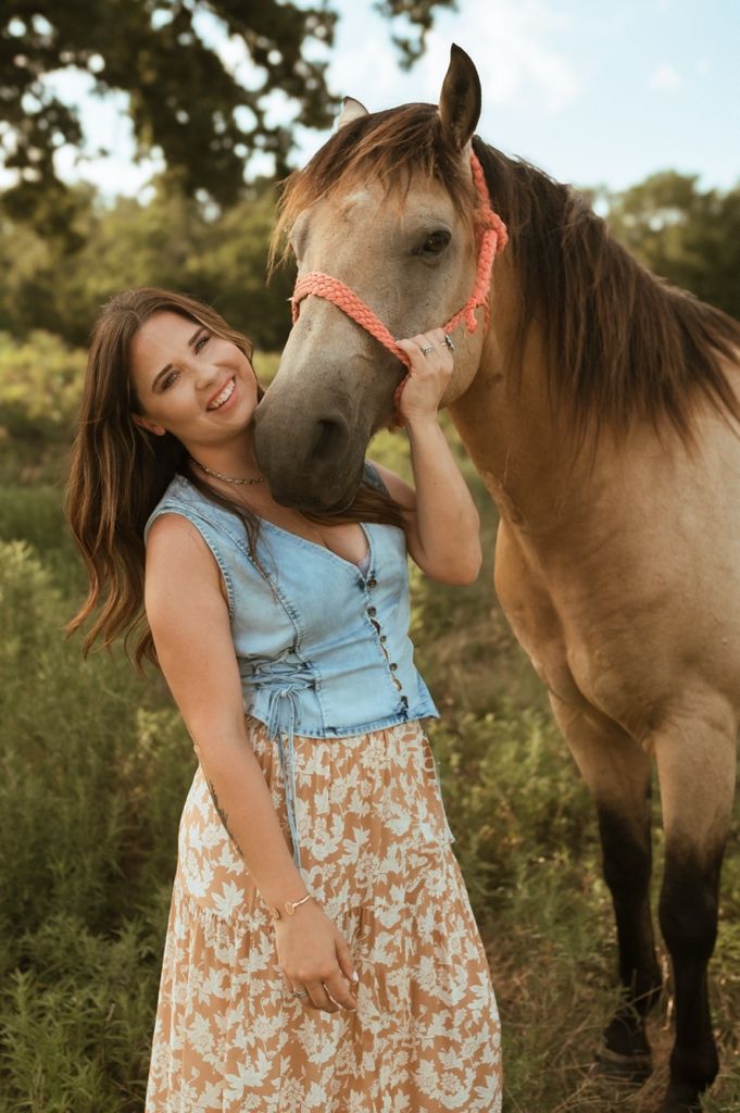 A beautiful buckskin horse wearing a pink halter stands with someone in a floral dress and denim top outdoors.