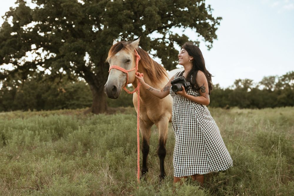 A gingham dress-clad figure stands in a grassy meadow petting a palomino horse at sunset.