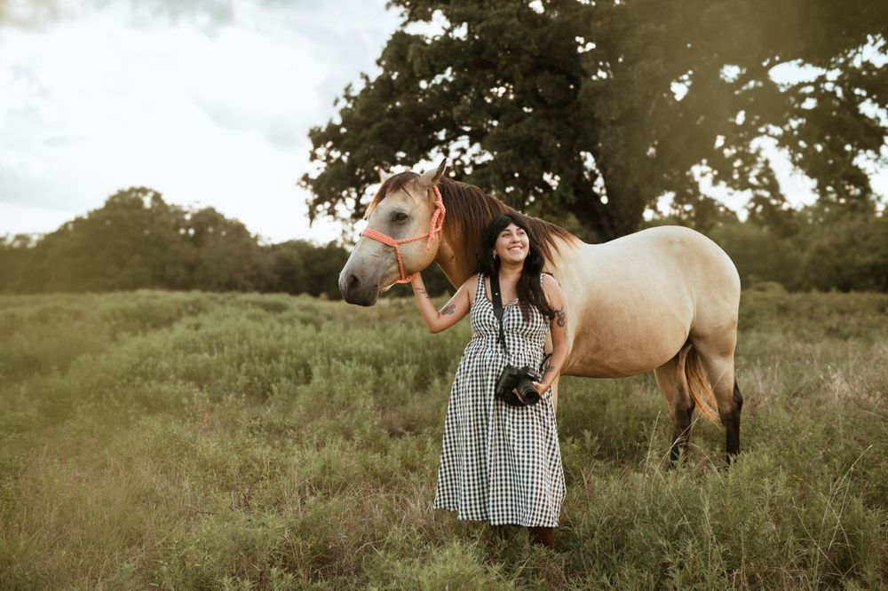 A woman in a gingham dress poses with a pale-colored horse in a sunlit meadow during golden hour.