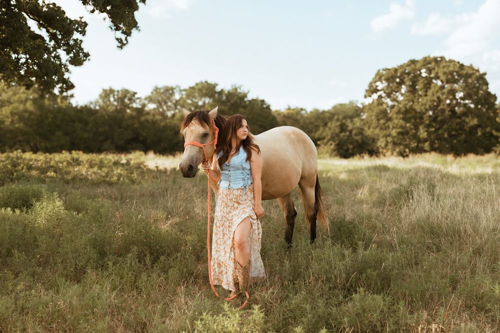 A floral maxi dress stands beside a tan horse in a sunlit meadow during golden hour.