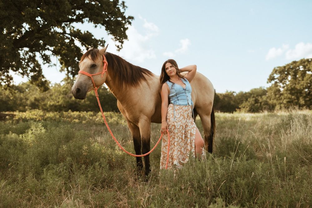 A brown horse and person in a floral skirt stand together in a sunlit meadow during golden hour.