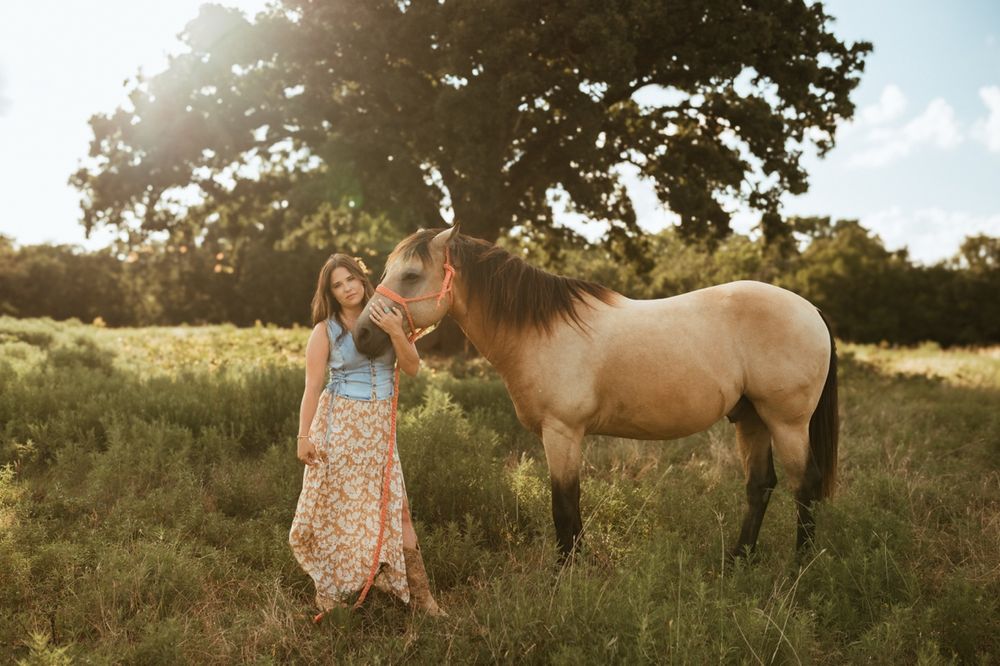 A dun-colored horse stands in a sunlit meadow beside someone in a floral dress during golden hour.