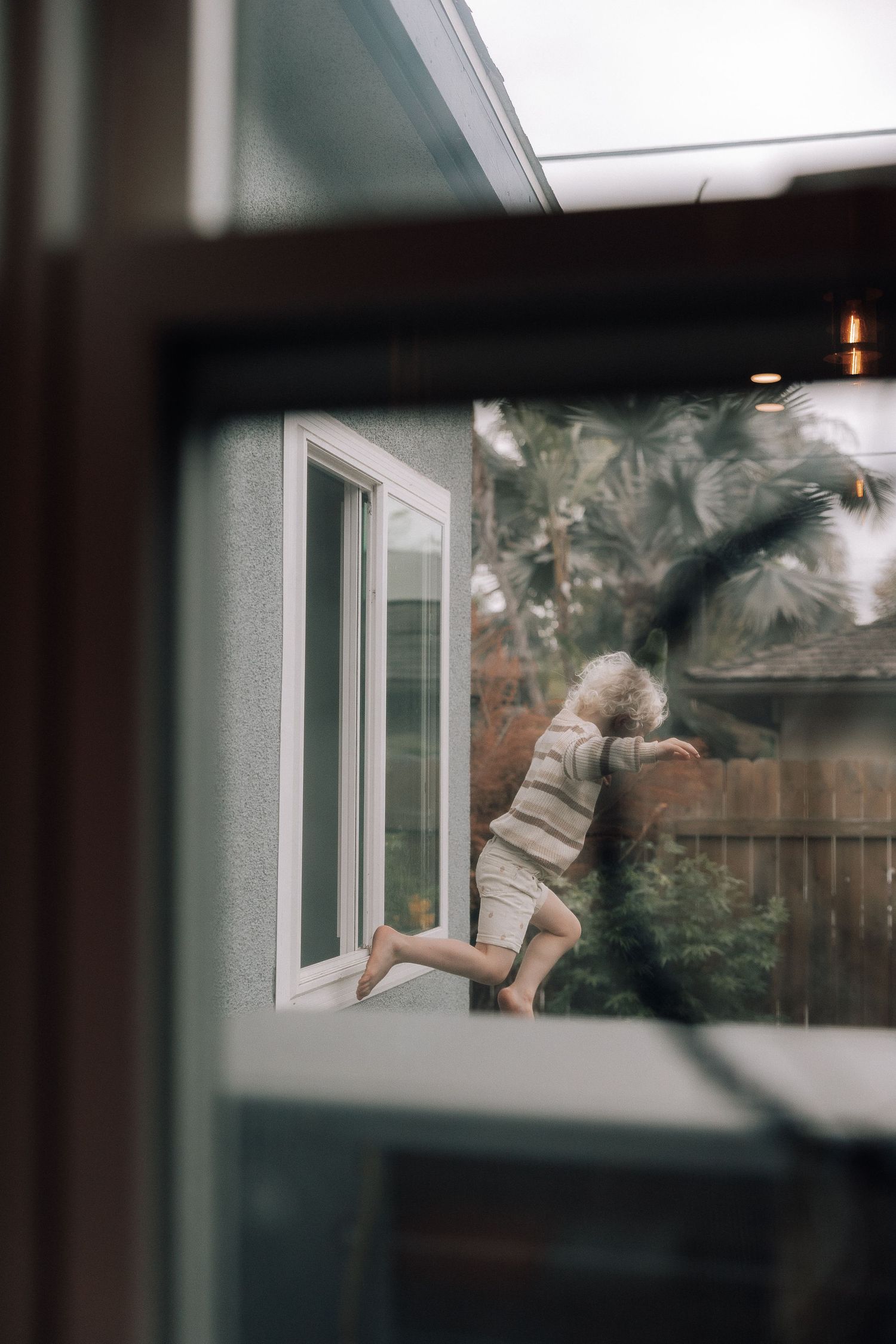 Through a window, a small child wearing a beige outfit climbs up the side of a house in a playful manner.