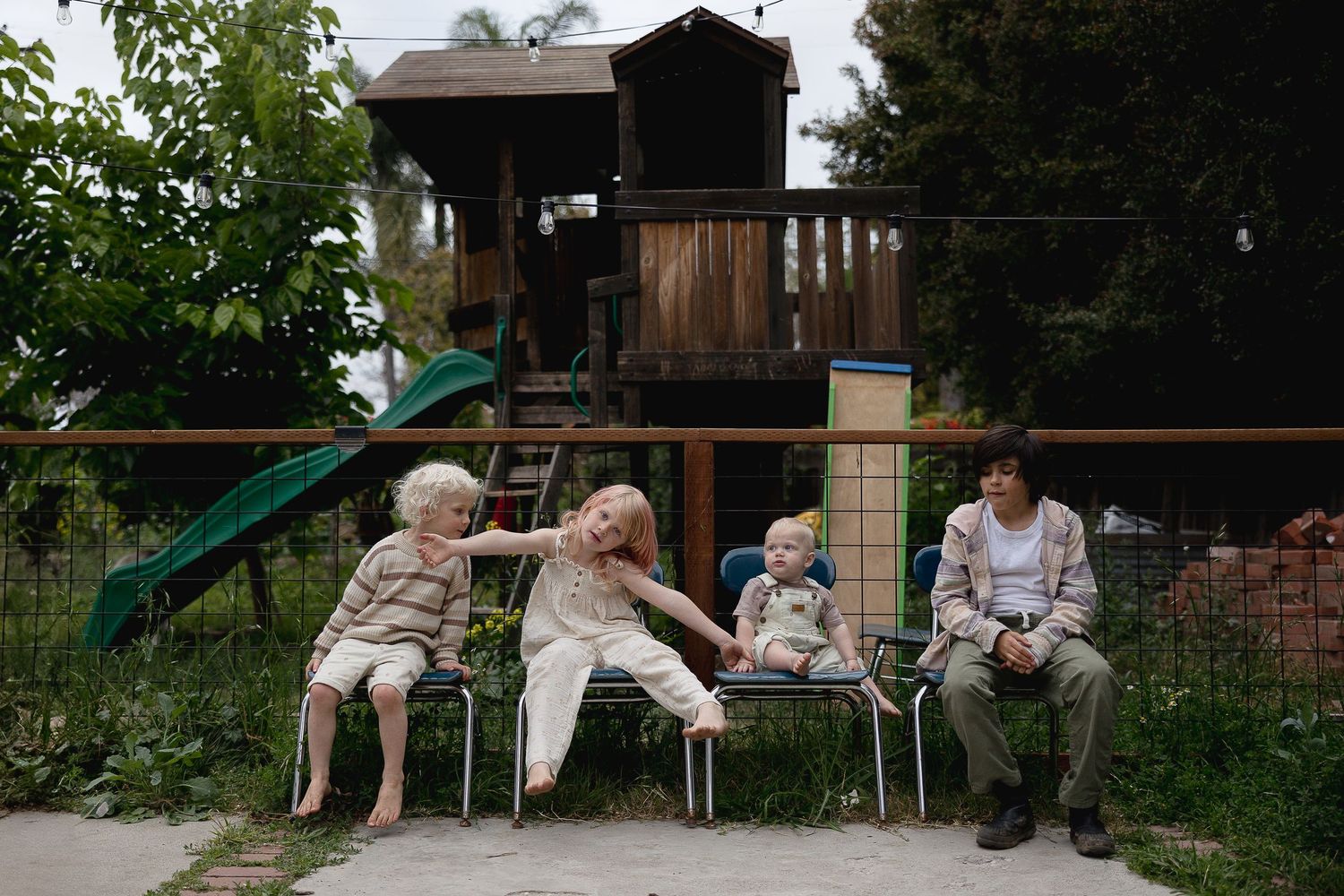 Four people sitting in white lawn chairs in front of a wooden playhouse in a backyard setting.
