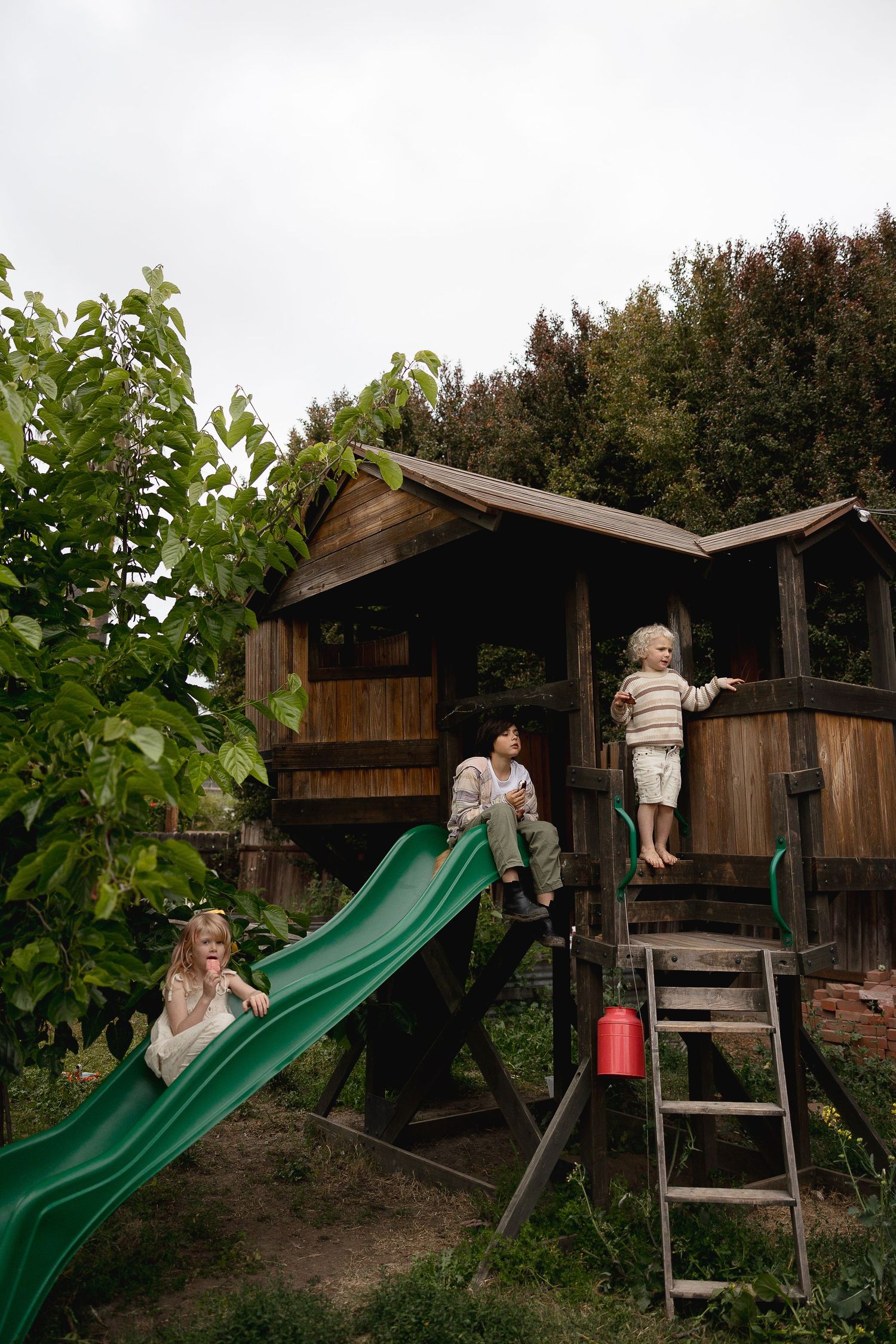 Children play on a brown wooden playhouse with a green slide in a backyard garden setting.