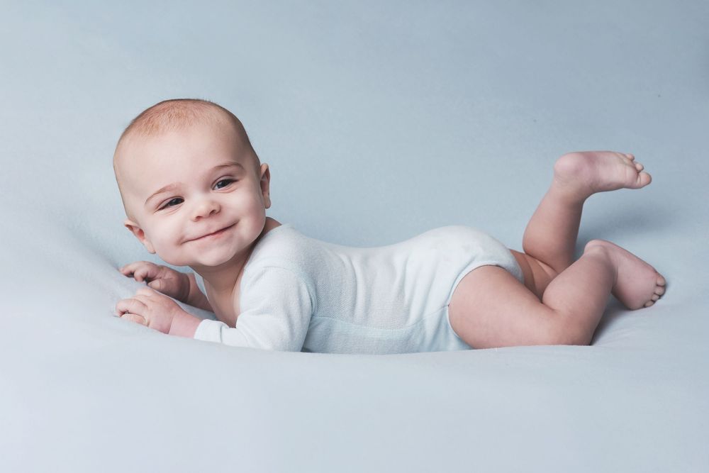 A young infant wearing a white onesie lies on their side on a light gray backdrop smiling.